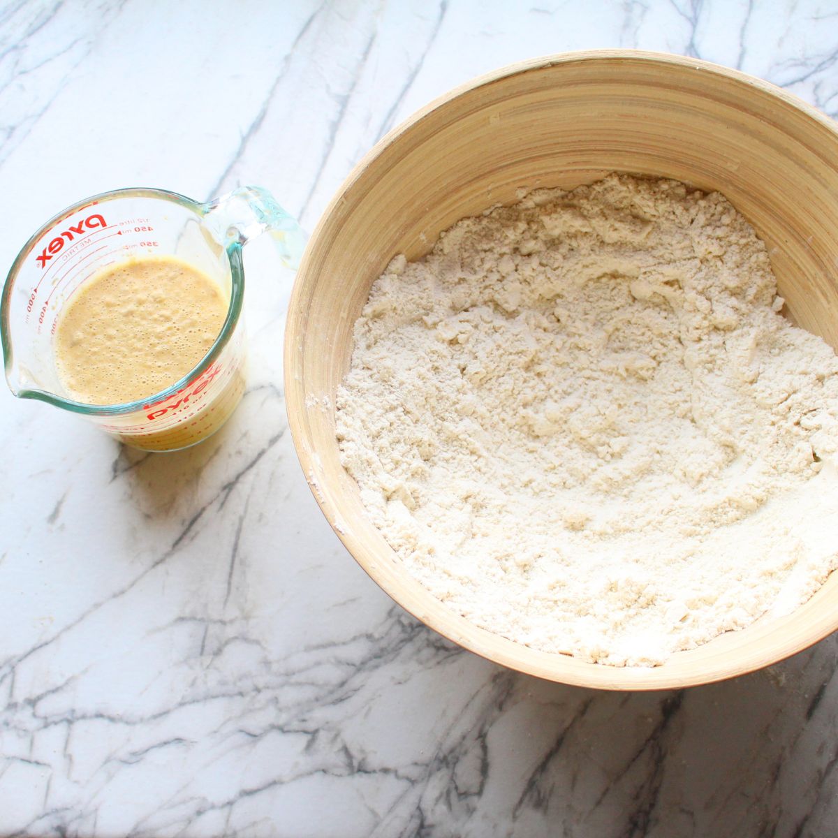 Liquid measuring cup on the left with liquid in it, and wooden bowl on the right with dry ingredients in it for making Swedish cardamom coffee cake.