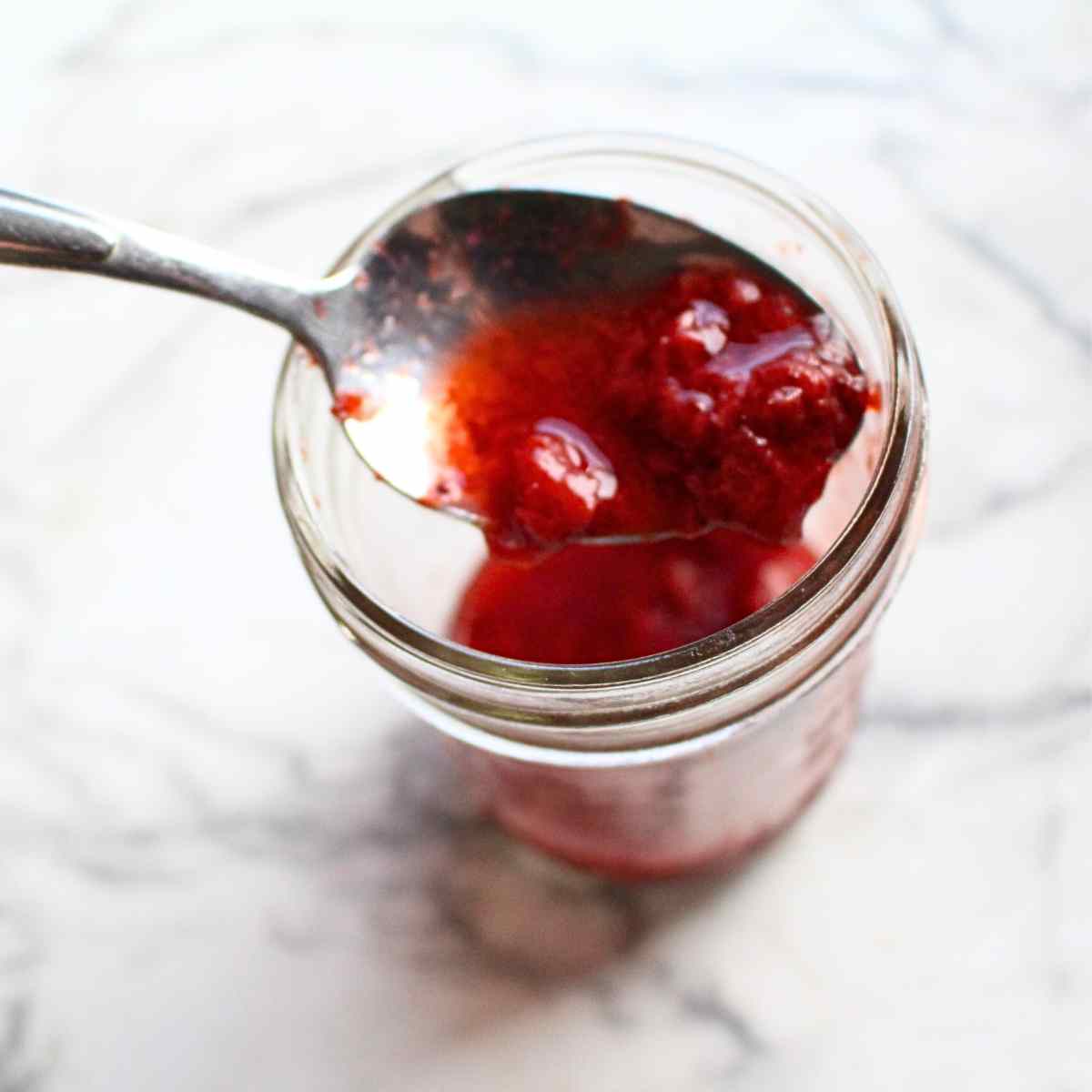 A spoon holding strawberry sauce with the small jar of strawberry sauce in the background.