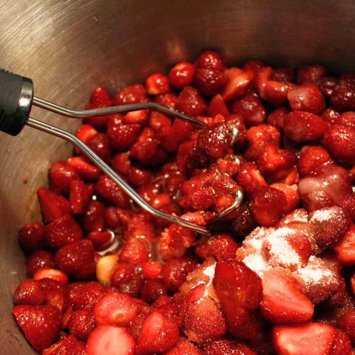 A potato masher in a pot starting to mash strawberries before they start cooking for strawberry sauce.