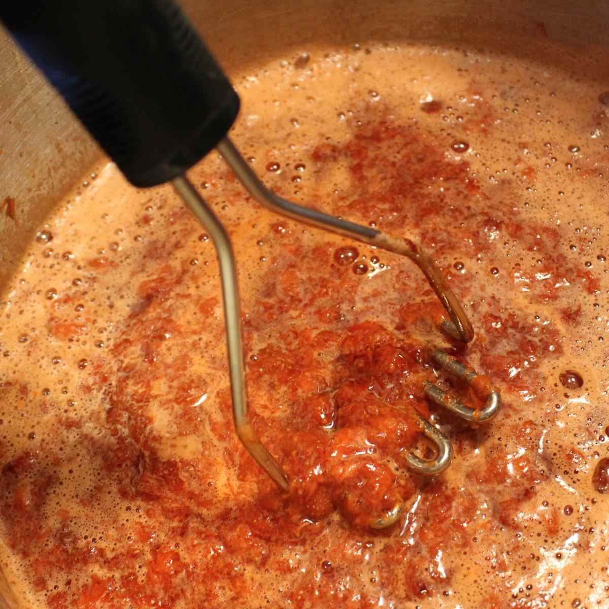 A potato masher in a pot mashing cooked strawberries for strawberry sauce.