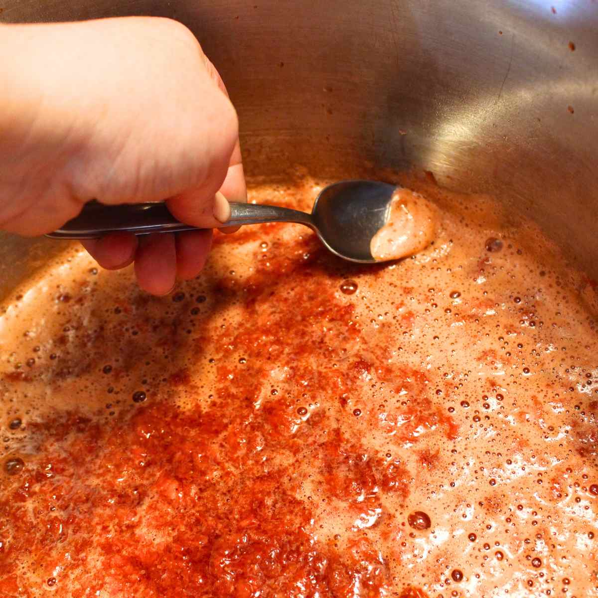 Showing a spoon skimming off the foam of strawberry sauce in a pot as it is cooking.