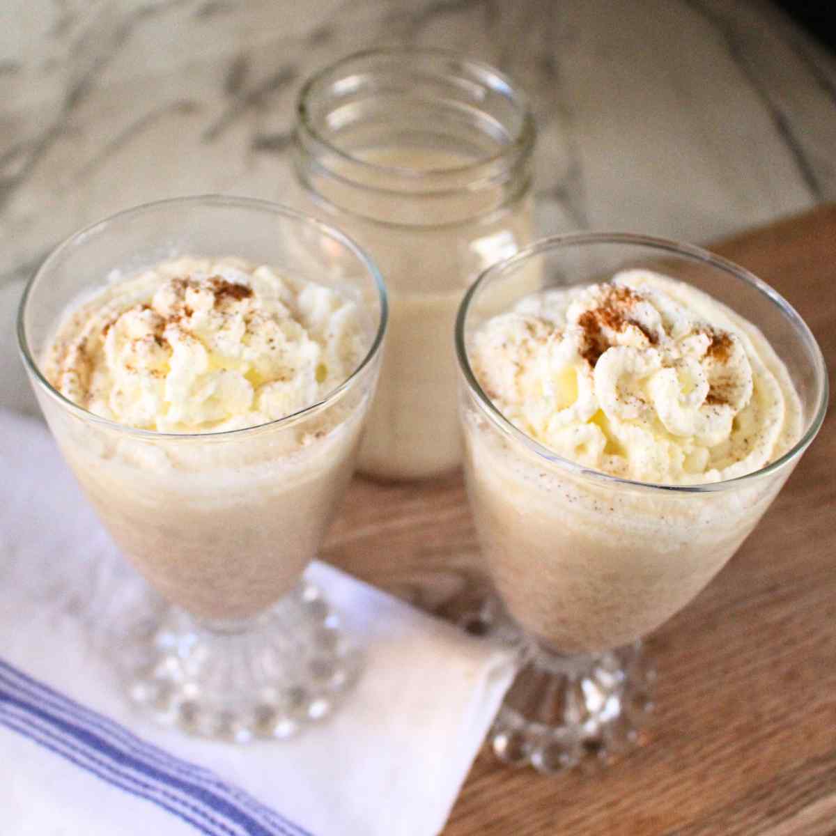 Two glasses of egg nog topped with whipped cream with a mason jar of more egg nog in the background.