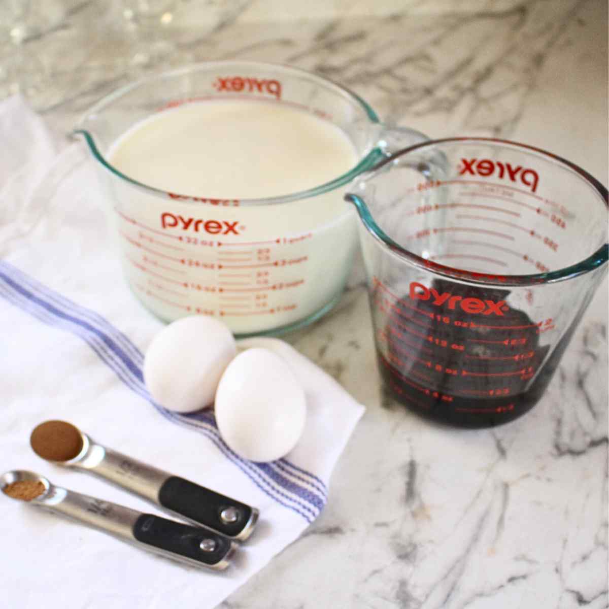 Ingredients for homemade egg nog including a measuring cup of milk, measuring cup with maple syrup, two eggs and two teaspoons of spices next to them.