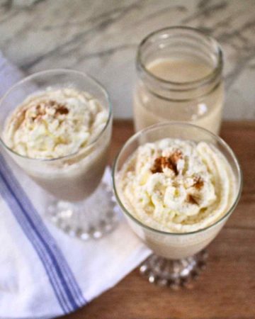 Two glasses of egg nog topped with whipped cream and a mason jar of more egg nog in the background.