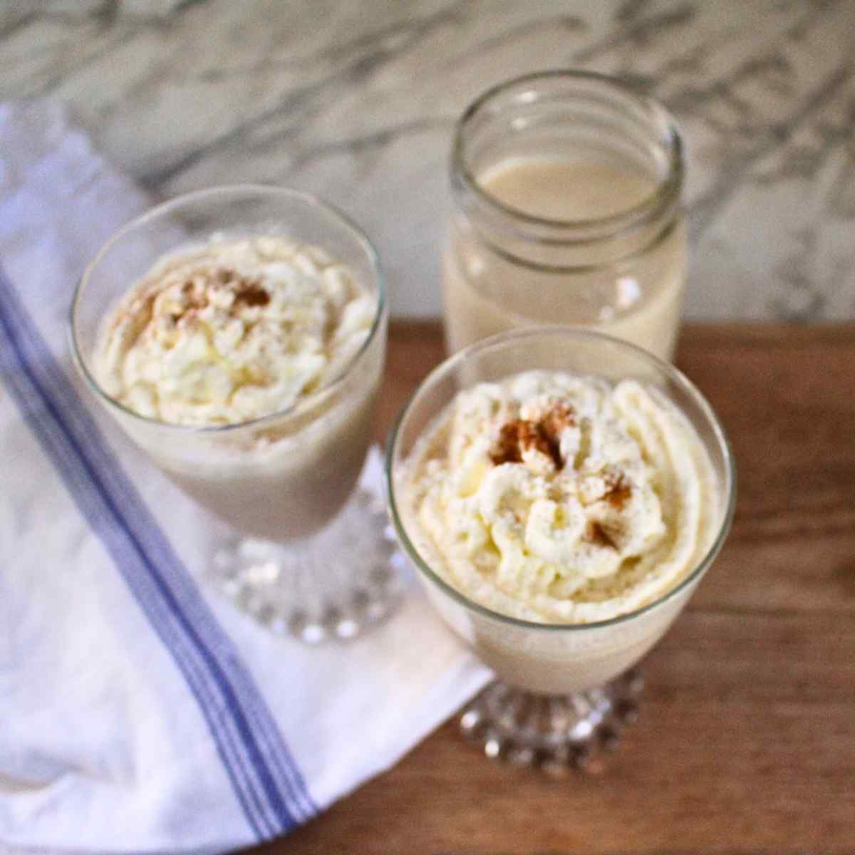 Two glasses of egg nog topped with whipped cream and a mason jar of more egg nog in the background.