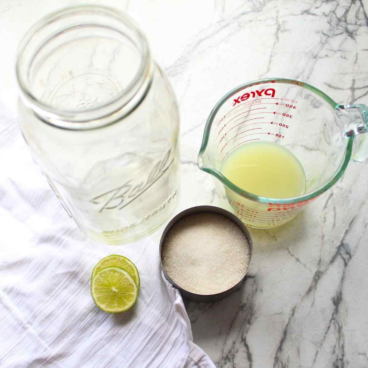 An empty mason jar next to a measuring cup with sugar in it, and a liquid measuring cup with lime juice in it for homemade strawberry limeade.