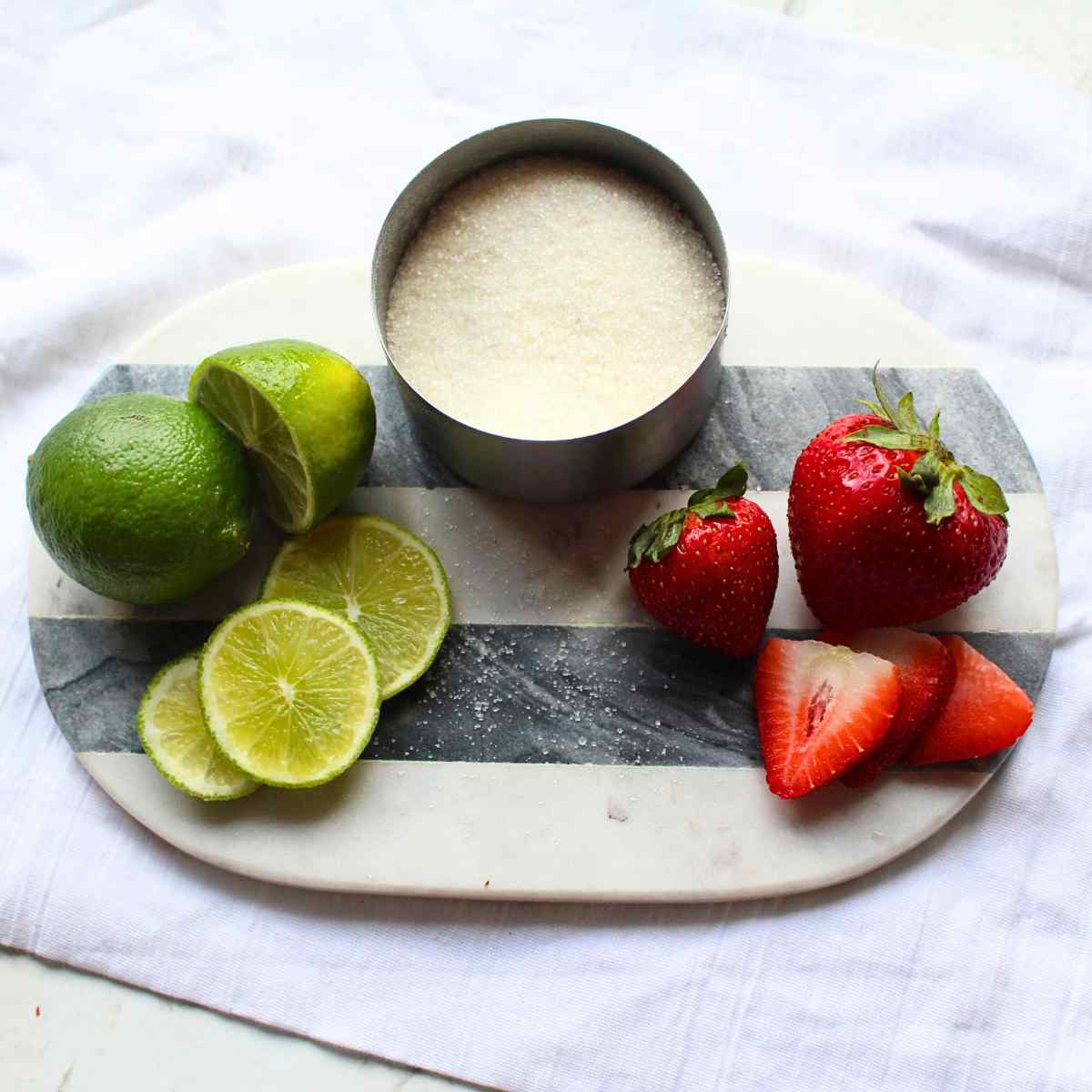 A small cutting board with limes on the left side, sugar in the middle, and strawberries on the right side.