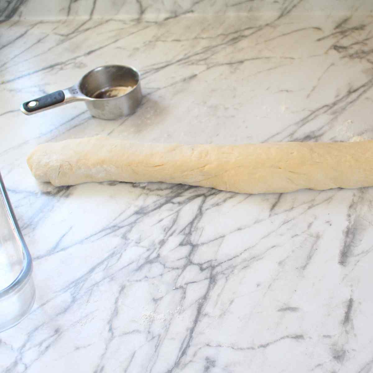 A rope of dough on a countertop with a cup of flour in the background.