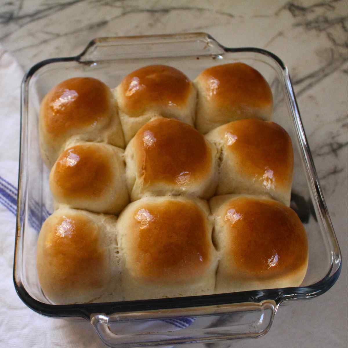 Baked and lightly brown dinner rolls in a baking pan, with melted butter spread on the top of them.