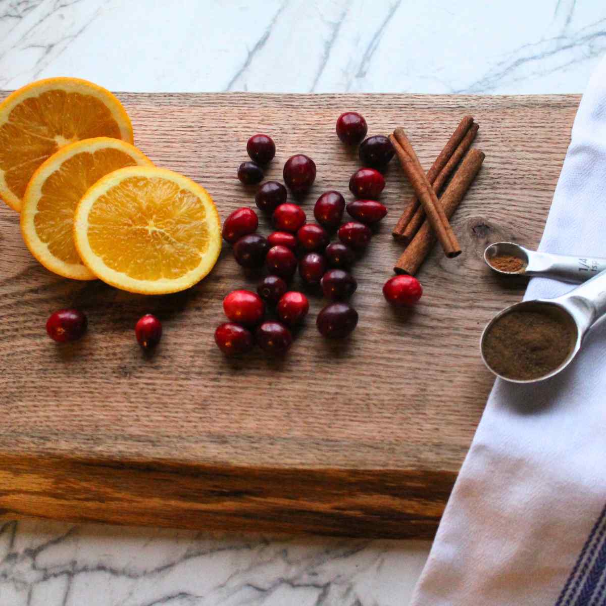 A cutting board with sliced oranges, cranberries, cinnamon sticks and measuring spoons of nutmeg and cloves on it for a Christmas simmer pot.