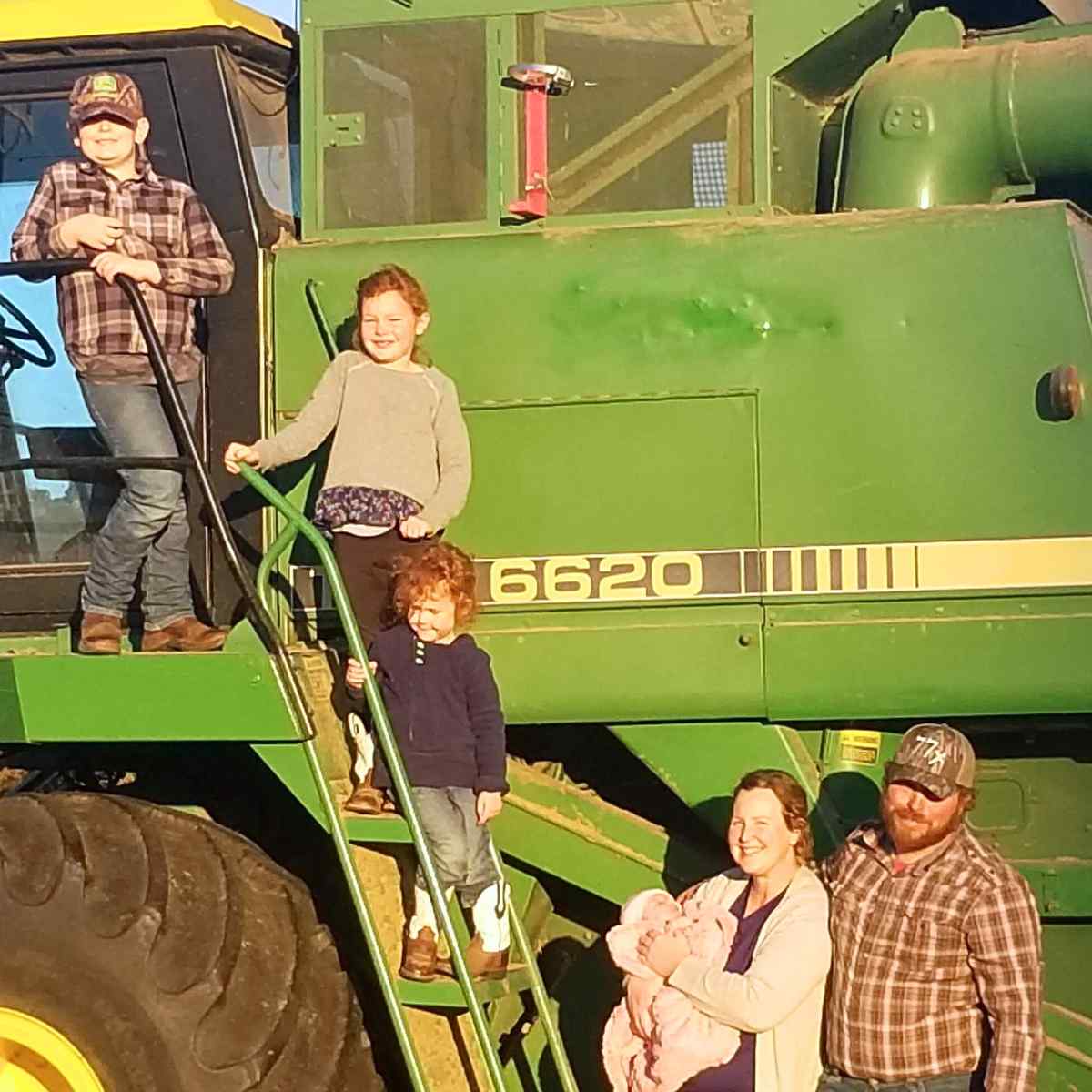 Three children on the railing of a combine, with a woman holding a baby and a man beside her on the ground next to the railing.