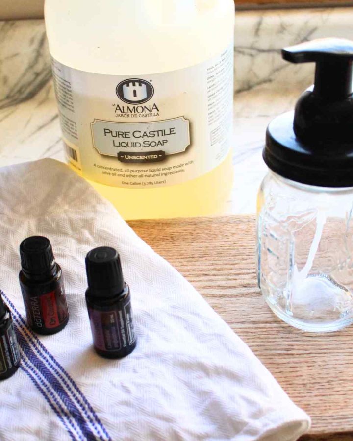 Three essential oil bottles on the left with a foaming hand soap bottle on the right with a jug of castile soap in the background.