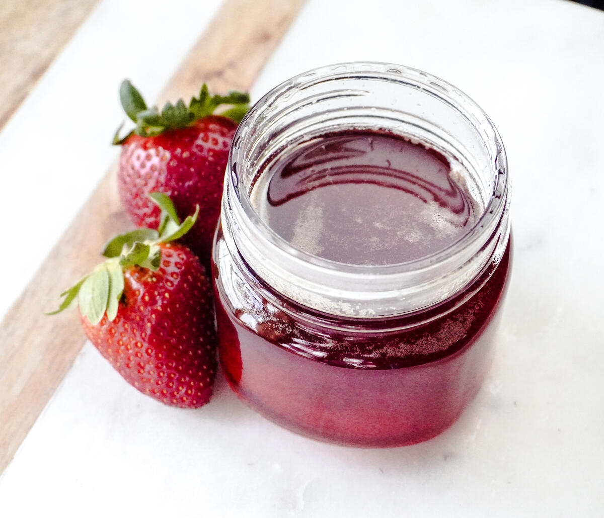 A jar of strawberry syrup with two strawberries next to it.